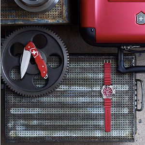 Rød Victorinox foldekniv foldet halvt ud. Vist ved siden af rødt ur og Victorinox kuffert. 